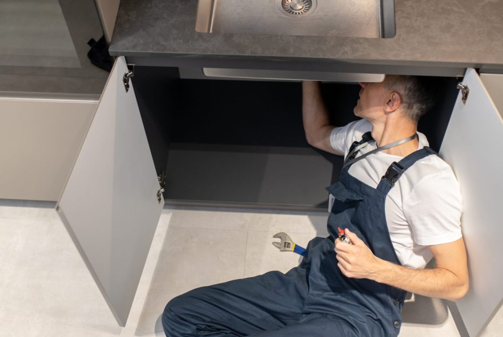 Solution. Top view of man plumber in overalls holding ball valve in hand lying on floor near open set of tools in kitchen fixing breakdown of sink